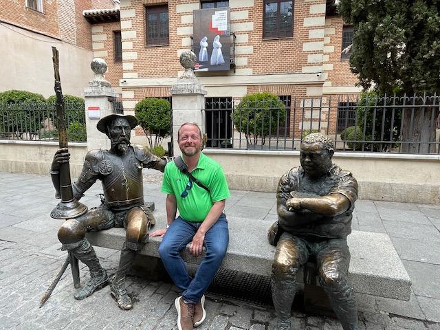 Josh Pope, 穿着绿色的polo衫和牛仔裤, 在马德里旅行时，他坐在桑丘·潘扎和堂吉诃德的青铜雕像之间, Spain. 在他身后是一座砖房，四周围着熟铁栅栏.