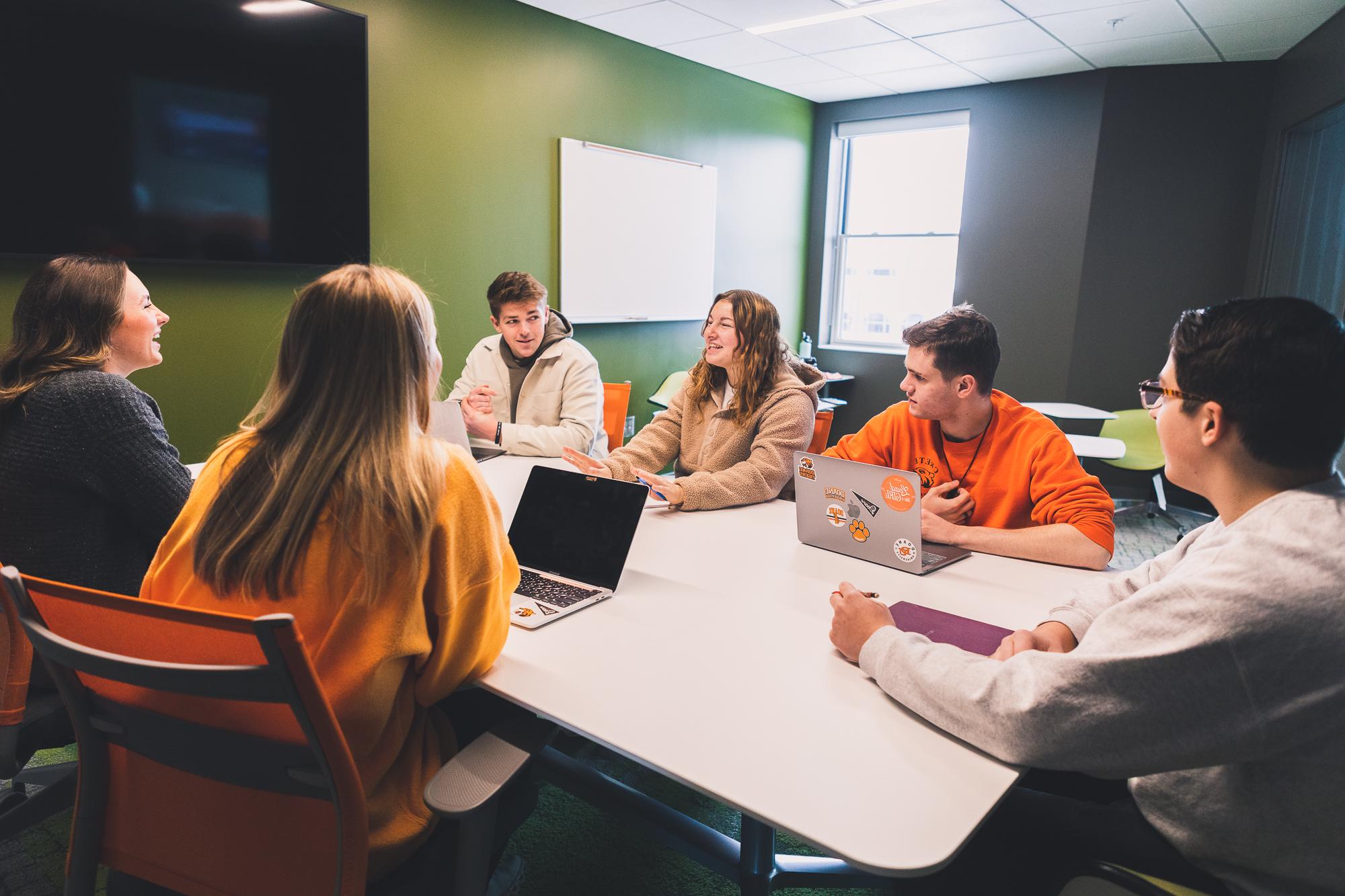Six students sit around a white, rectangular table in a vibrant green-painted study room. Behind them are individual desks and chairs, and a window. 
