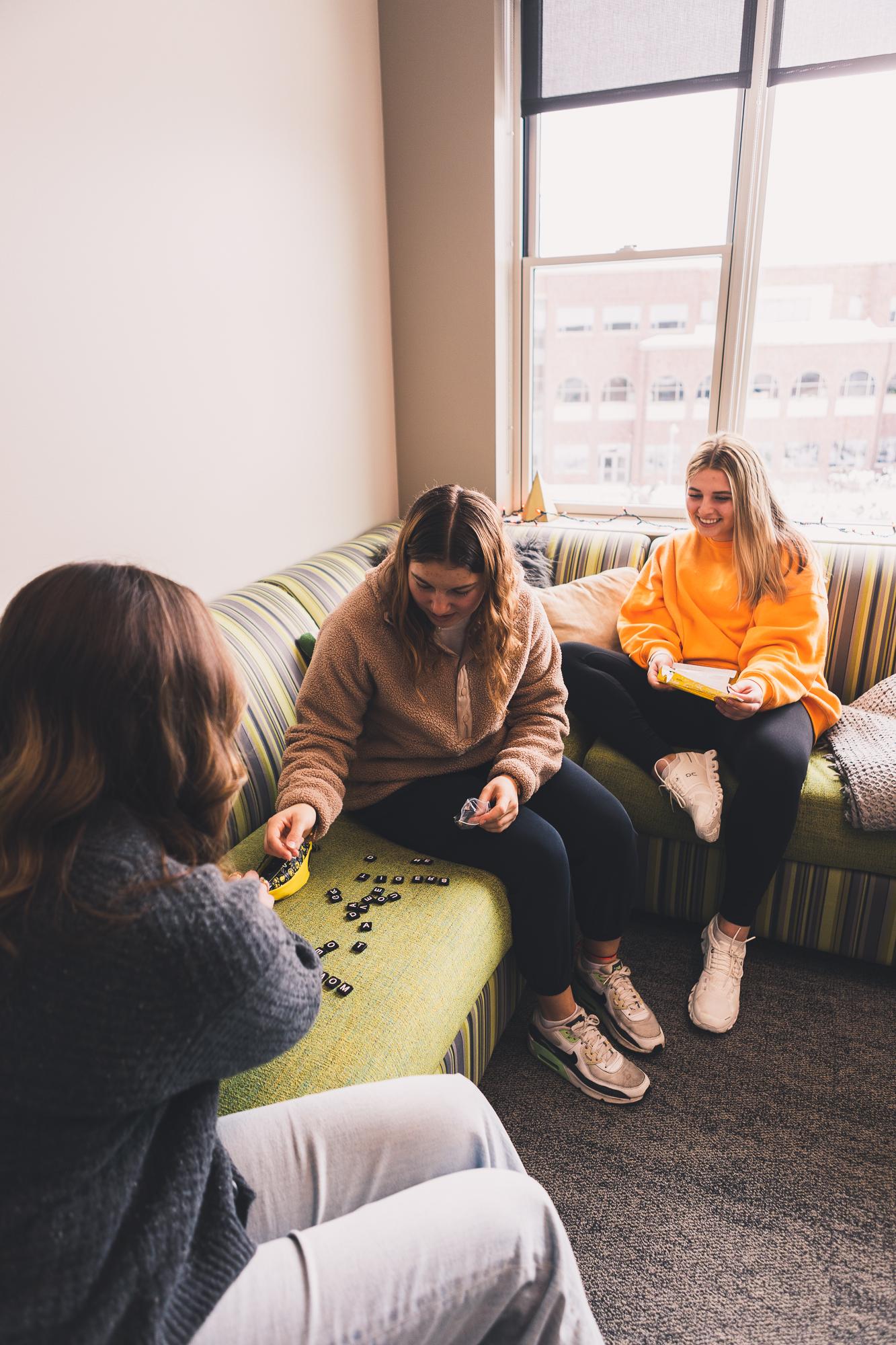 Maci McCarter reads from a pamphlet of directions while Jaiden Papik and Grace Schroller play Bananagrams on a green couch in a third-floor suite. Behind them is a large window showing views of Doane's Art/Ed building.