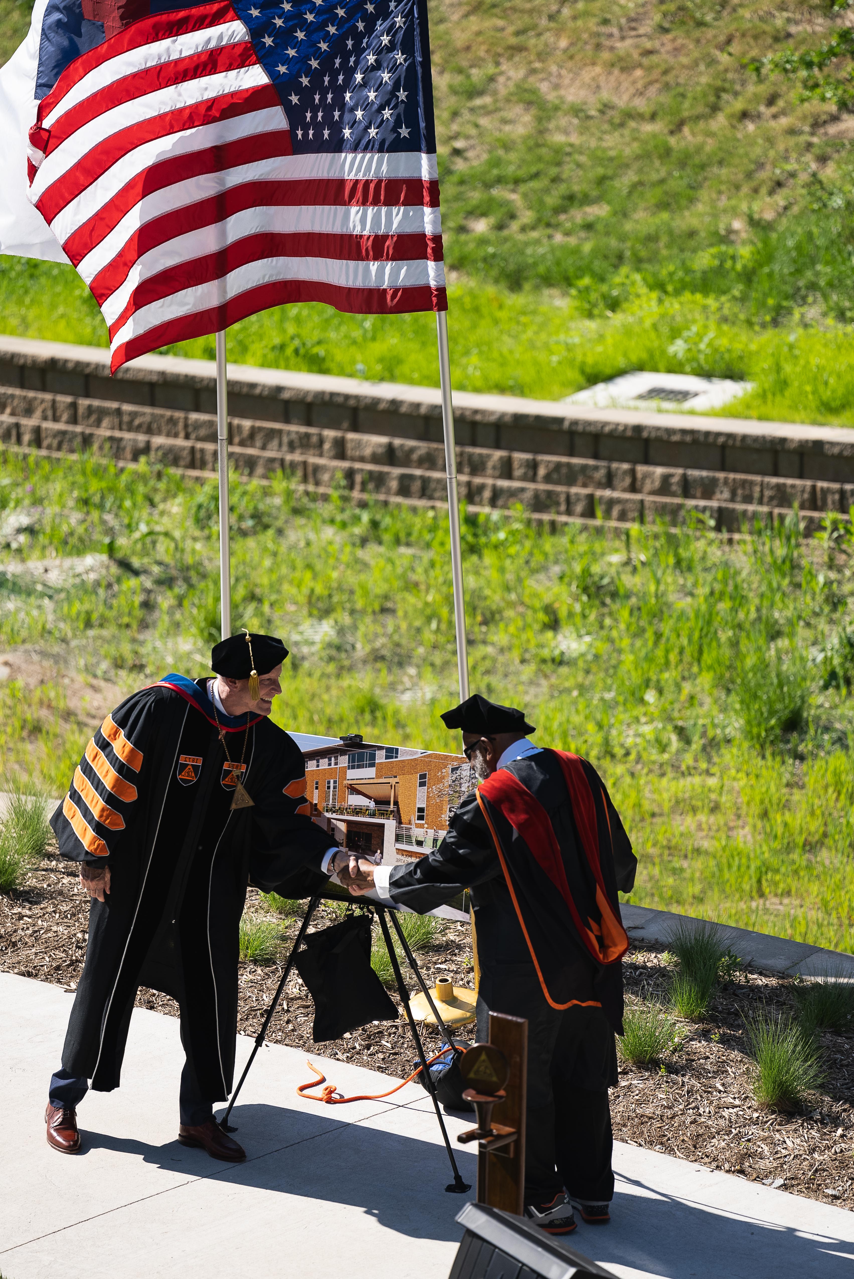 两名身穿黑色学术袍的男子在一幅建筑海报前握手. 他们身后飘扬着一面美国国旗, 绿色植物生长的背景被砖挡土墙隔开.