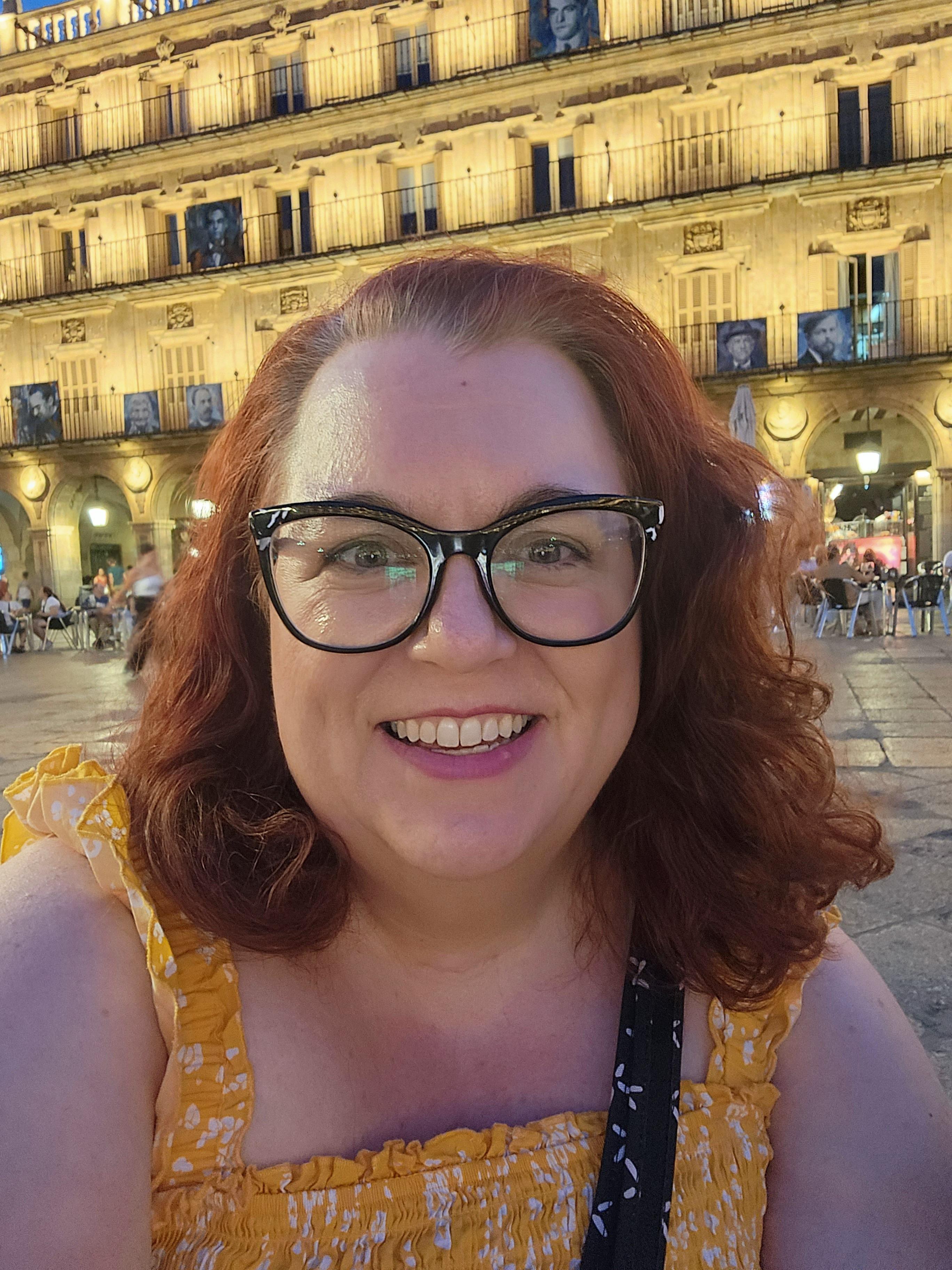 梅勒妮·达米科在西班牙旅行时拍了一张自画像. 她有一头卷曲的红头发，戴着一副大大的猫眼眼镜，穿着一件黄色的上衣.