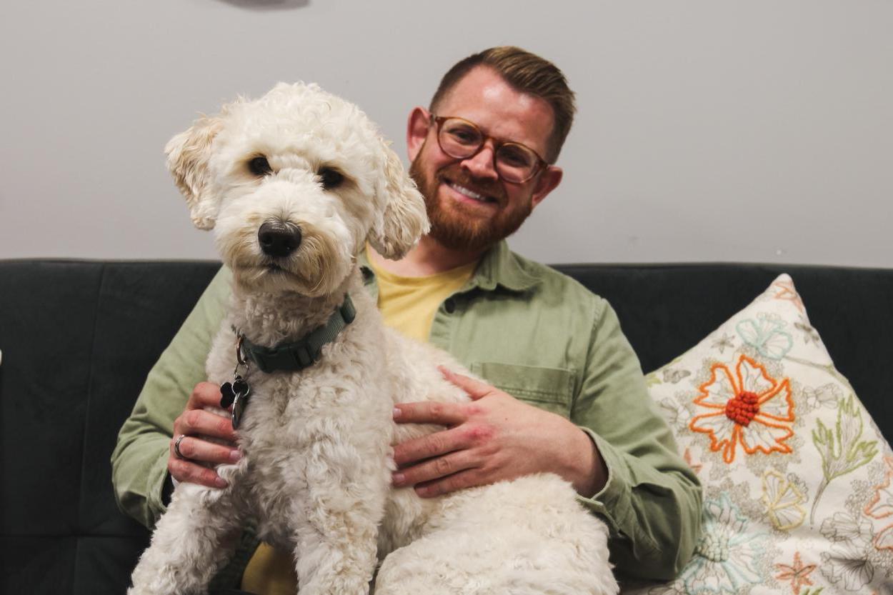 Georgie, a light-colored goldendoodle with fluffy trimmed fur, sits on the lap of his person, Blake Tobey, academic specialist in the Academic Success Center. The two sit on a comfy grey couch in the center's office in the Communications Building. 