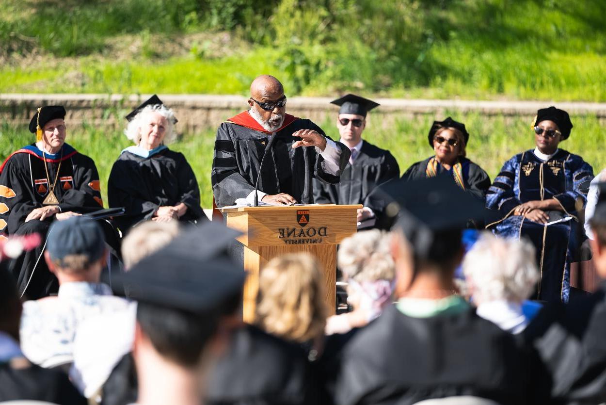 一名身穿学术袍的黑人男子在讲台上向身穿黑色长袍、戴着毕业帽的听众做手势. 在他身后是五个人，也穿着学术袍.