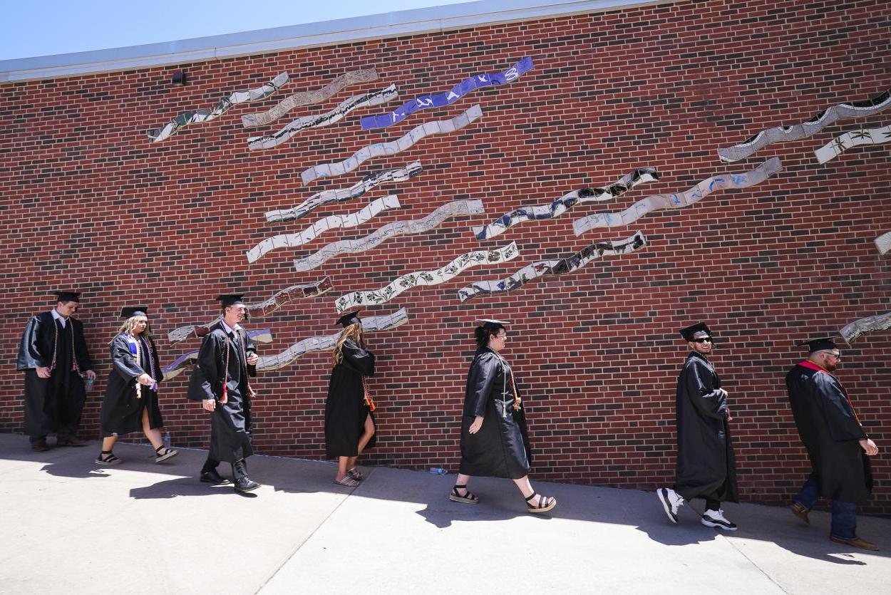 七名戴着黑色帽子、身穿黑色长袍的学生走下斜坡，前方是一面砖墙，墙上是一幅由波浪瓷砖制成的壁画.
