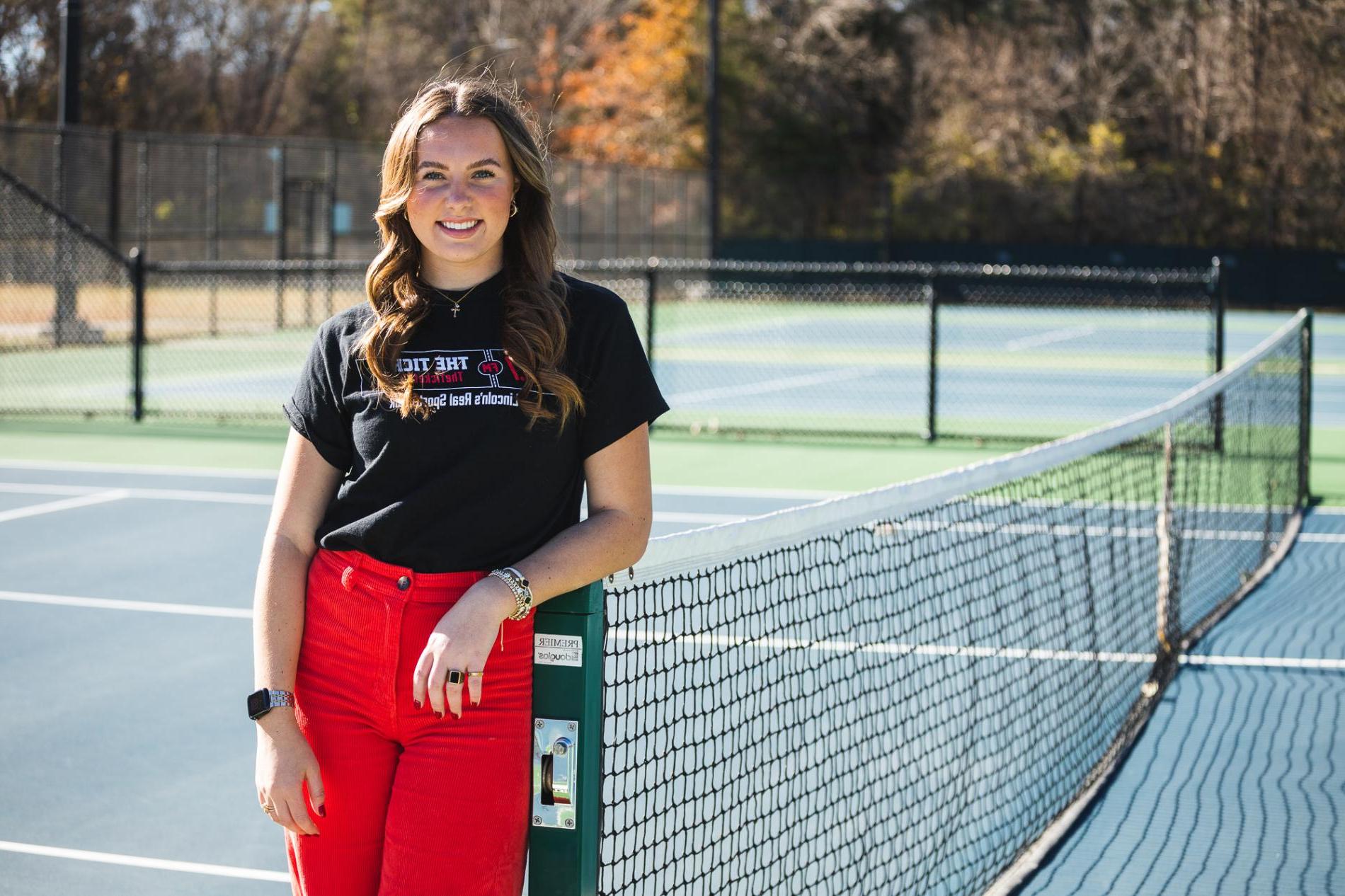 恩典Schroller leans against a green pole holding up a tennis net on Doane's blue-painted courts. She wears a black t-shirt with the logo for "The Ticket" and red corduroy pants.