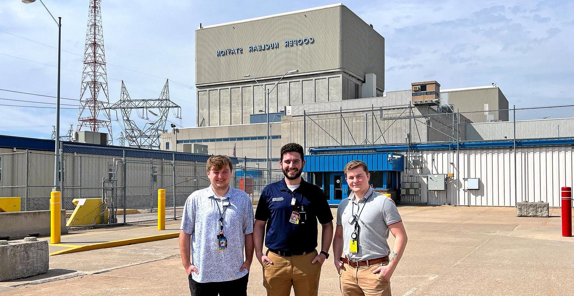 Three men in polos with badges stand on a concrete parking lot in front of a blocky concrete building bearing the words "Cooper Nuclear Station."