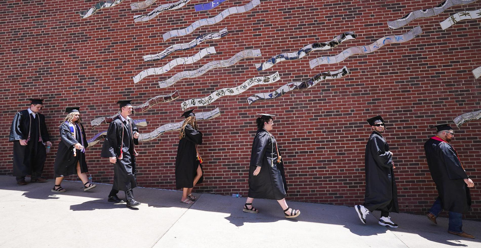 七名戴着黑色帽子、身穿黑色长袍的学生走下斜坡，前方是一面砖墙，墙上是一幅由波浪瓷砖制成的壁画.