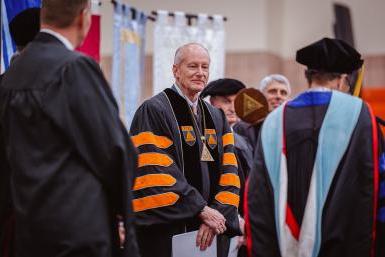 Dr. Roger Hughes stands on a stage in black and orange robes as the newly inaugurated 13th President of Doane University.
