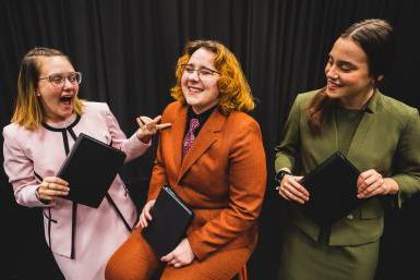 Students Maddy Ramey, wearing a green suit; Olivia Vore, wearing an orange suit with pink tie; and Ali Moulton, wearing a pink suit, laugh in front of a dark curtain while holding small black binders, used for interpretive events in competitive speech.