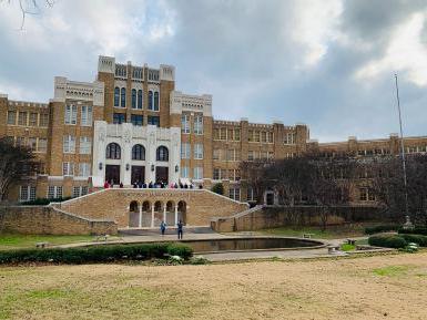 An image of the front of Central High School, in Little Rock, Arkansas. The school is yellow brick with a reflecting pool and grass lawn in front. Tourists mill around the pond and up stairs to the doors of the school.