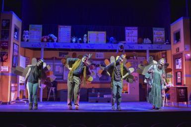 Four actors on a stage lit with purple and yellow lights, a woman in a long dress, two men wearing dark pants, shirts and fringed vests, and a second woman in a tshirt, leggings and boots. They're all wearing felt turkey tails and leaning to their right. The stage is set to look like a theater classroom, with props and posters on three walls and the fourth open to the audience.