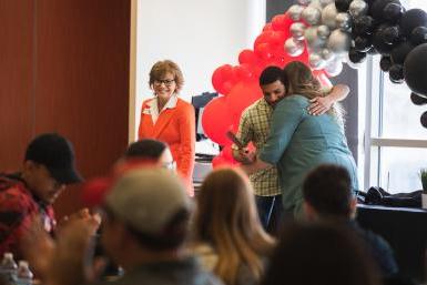 Senior Arian Alai, wearing a green plaid shirt, hugs Jill Kline, wearing a teal jacket, in front of a black, silver and red balloon arch. 在前景，学生和他们的家人坐着，只有阿里安和吉尔在焦点上.