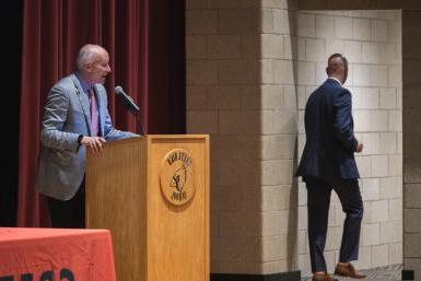 Doane President Dr. Roger Hughes addresses employees of Crete Public Schools behind a wooden podium with the Crete High School cardinal head logo. CPS Superintendent Dr. Josh McDowell walks offstage through a doorway. Behind Hughes is a red curtain. 