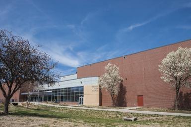 Image of the exterior of Seward High School, a red brick building with an entrance made of glass windows and doors, and light grey and stone-colored blocks. Trees in front are blooming and the grass is turning green. The sky overhead is bright blue with wispy clouds. 