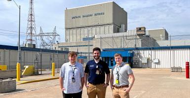 三名身穿polo衫、戴着徽章的男子站在混凝土停车场上，前面是一栋混凝土建筑，上面写着“库珀核电站”."