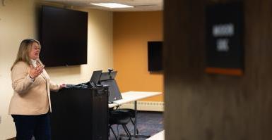 Dr. Jen Bossard speaks at the front of the College of Business's technology classroom, located in the Fred Brown Center in Lincoln. An open door partially covers the image, and behind Bossard are two large television screens, a tablet and a tech cabinet.
